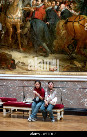 Two oriental women sit on a red velvet bench under a giant canvas depicting French King Henry IV entering Paris Stock Photo