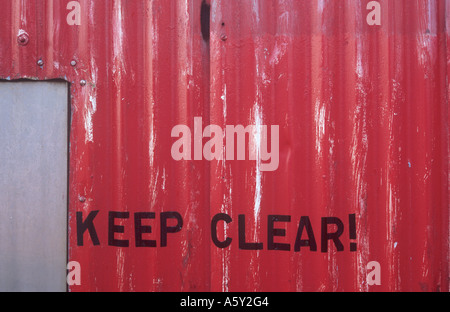 Detail of a red painted corrugated iron wall with an aluminium door set in it and proclaiming Keep clear! Stock Photo