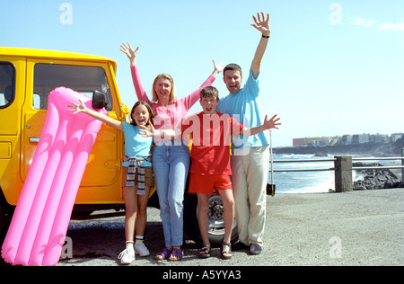 HOLIDAY FUN SNAP PHOTO EXCITED FAMILY Colourful happy young family of four with rental hire car on holiday wave and pose for the camera by the sea Stock Photo