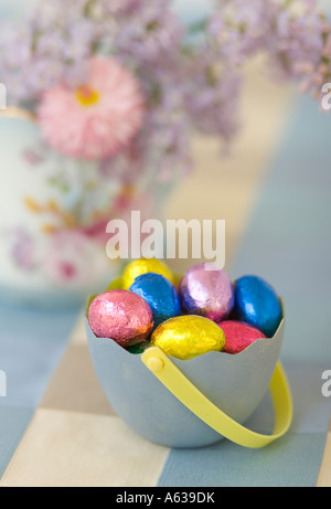 Foil-covered chocolate Easter eggs in an Easter container, with soft-focus flower arrangement in background. Stock Photo