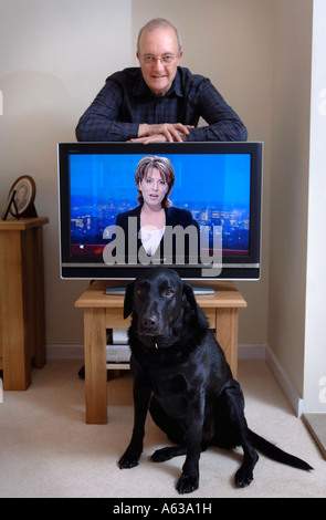 A MIDDLE AGED MAN LEANS ON A TELEVISION WHILE A PET BLACK LABRADOR SITS ON THE FLOOR UK Stock Photo