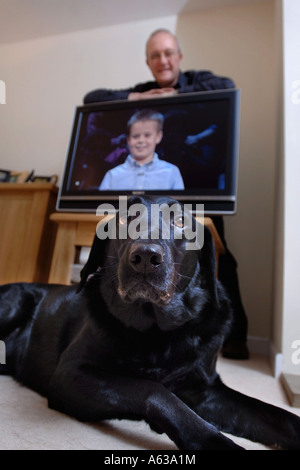 A MIDDLE AGED MAN LEANS ON A TELEVISION WHILE A PET BLACK LABRADOR LIES ON THE FLOOR UK Stock Photo