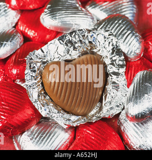 A single milk chocolate heart unwrapped from its foil, on other hearts wrapped in red and silver foil. Stock Photo