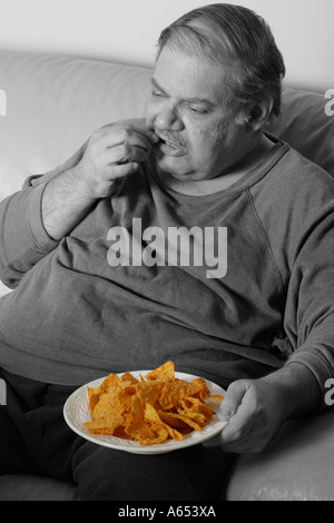 Overweight man eats chips while sitting on his couch at home. Stock Photo