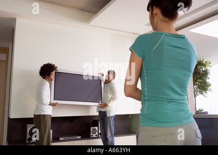 Young woman watching as two young men move plasma television, back view Stock Photo