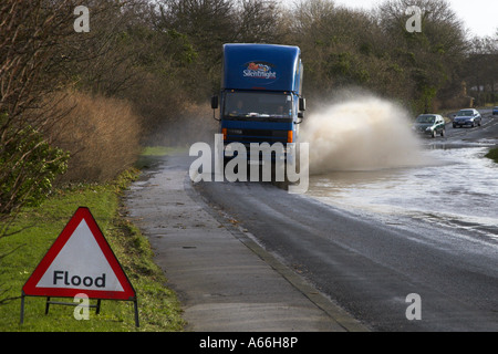 Large lorry driving (cars vehicles behind) splashing through flood water on rural road (warning sign in foreground) - near Otley, West Yorkshire, UK. Stock Photo