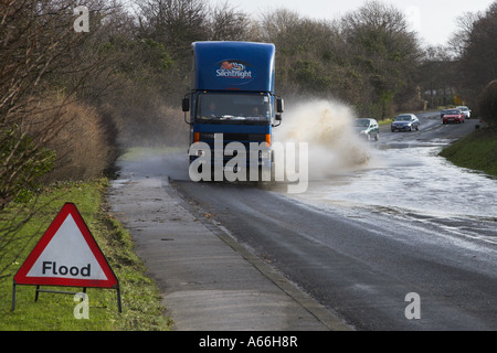 Large lorry driving (cars vehicles behind) splashing through flood water on rural road (warning sign in foreground) - near Otley, West Yorkshire, UK. Stock Photo