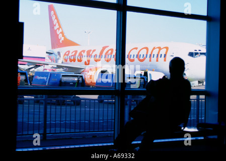 Man seated in airport departure lounge looking at aeroplane parked on stand through window