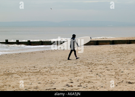 Surfer carrying his surfboard across the beach dressed in his winter wetsuit at the end of a dull winter day Stock Photo