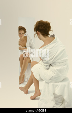 Pretty young woman in white robe inspecting herself in full length mirror in studio Stock Photo