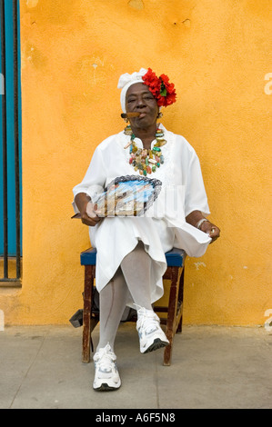 A Cuban woman sitting in the sunshine in front of a pastel pink painted wall smoking a long Cuban cigar, Havana, Cuba Stock Photo