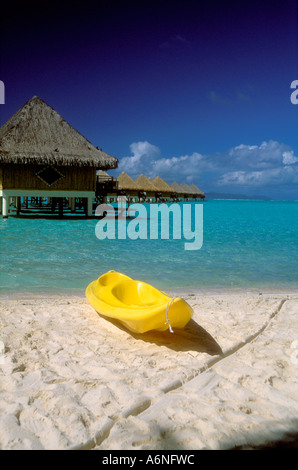 Yellow canoe / kayak on beach with thatched roof bungalows over lagoon Stock Photo