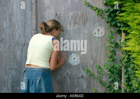 Teenage girl peeking through hole in fence Stock Photo