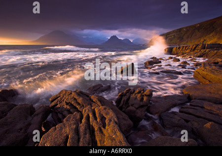 A shard of golden light shines brightly on the Cuillins on the horizon.  The incoming tide pounds the foreshore at Elgol Stock Photo