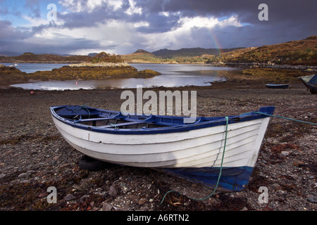 A wooden rowing boat lies stranded on a beach near Plocton, Scotland awaiting the arrival of high tide.  A rainbow is overhead Stock Photo