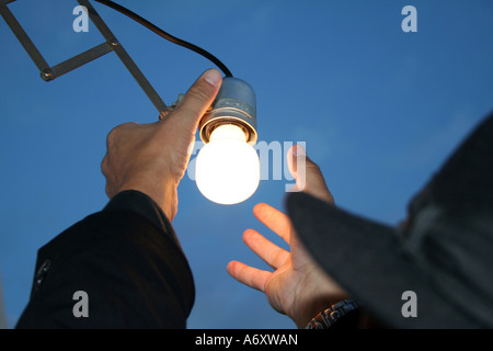 Hands replacing an illuminated light bulb in a socket against a blue evening sky, Cagliari, Sardinia, Italy. Stock Photo