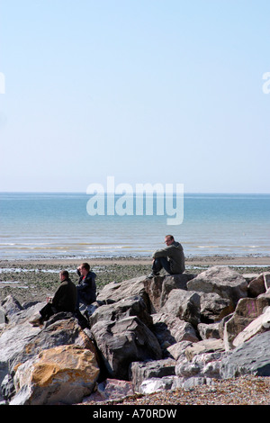 Three young men sitting on rocks on beach at Goring-by-Sea, West Sussex, UK Stock Photo