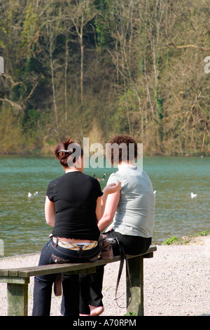 Two young women sitting by edge of lake smoking cigarettes Stock Photo