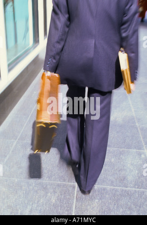 Low section, rear view of businessman walking away, in business attire and with a briefcase and papers under his arm, on the street. Stock Photo