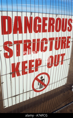 White sign behind wire mesh barrier with symbol banning people and large red letters stating Dangerous structure Keep out Stock Photo
