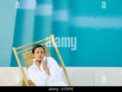 Woman lying in lounge chair at edge of pool, wearing bathrobe Stock Photo