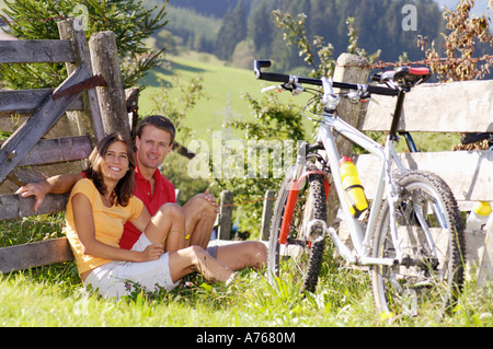 Young couple sitting in meadow with mountain bike, leaning on wooden fence Stock Photo