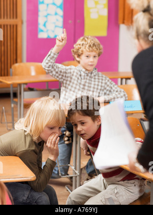 Boy (4-7) raising hand in classroom, focus on boys whispering in foreground Stock Photo