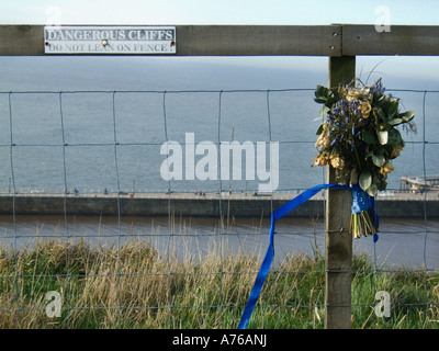 Dangerous Cliffs Do Not Lean on Fence sign on a cliff top wire mesh fence, alongside a dead bunch of flowers tied to a fence post with a blue ribbon Stock Photo