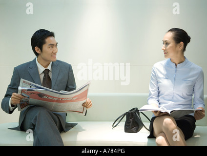 Businessman and woman sitting on waiting room bench, smiling at each other Stock Photo