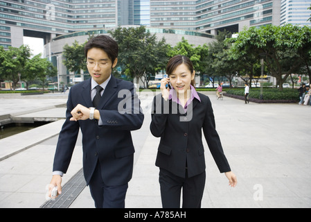 Two young executives walking in office park, one checking watch while other uses cell phone Stock Photo