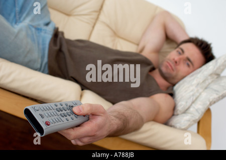 man laying on a sofa watching television changing channels with a TV remote zapper Stock Photo