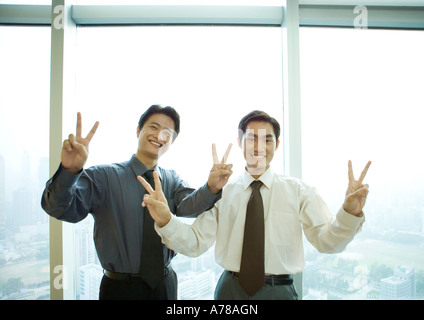 Two businessmen standing near window in high rise, making victory sign, smiling at camera Stock Photo