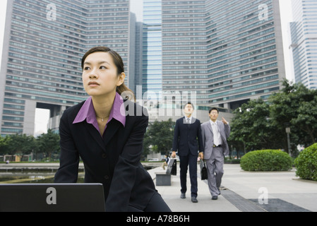 Businesswoman using laptop in office park, two businessmen approaching in background Stock Photo