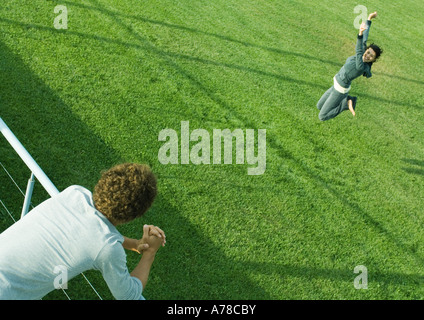 Teenage boy leaning on railing, watching teenage girl jumping on grass Stock Photo