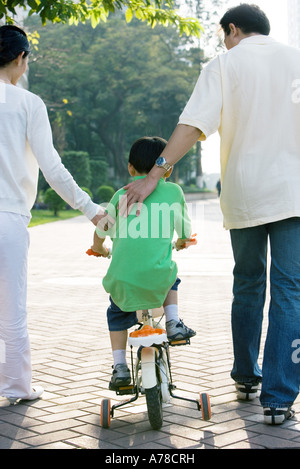 Boy riding bicycle with training wheels, parents on either side, rear view Stock Photo