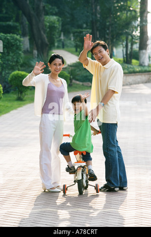 Boy riding bicycle, parents on either side, waving at camera Stock Photo