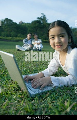 Girl lying in grass using laptop, parents sitting in grass in background Stock Photo
