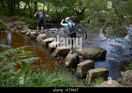 Mountain Bikers Carrying Bikes across Stepping Stones on Dartmoor. Stock Photo