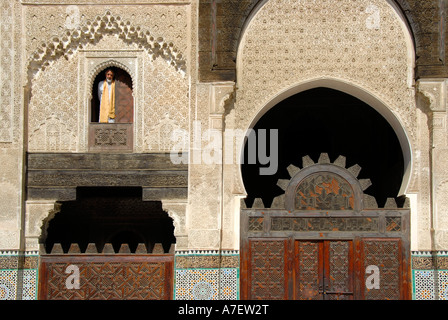 Man wearing traditional cloth looks out of a window decorated with rich ornaments above doors and gates Medersa Bou Inania Fes  Stock Photo