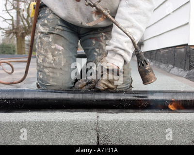 Roofer on knees using gas flame to make bitumen backing stick on 'flat' garage roof felt renewal (replacing old felt) England UK Stock Photo