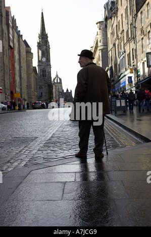 old elderly man in long coat hat and with a walking stick on wet pavement with reflection the royal mile Stock Photo