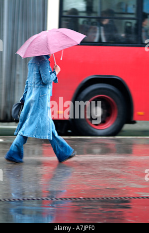 Woman Walking Along a City Pavement In Front of a Red Bus During a Rain Shower Stock Photo