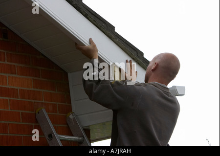 shaven headed workman aligning and installing white pvc cladding over old wooden panels on a red brick bungalow Stock Photo