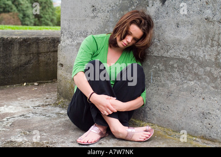 A depressed young woman sits leaning against a concrete wall  hugging her knees. Stock Photo