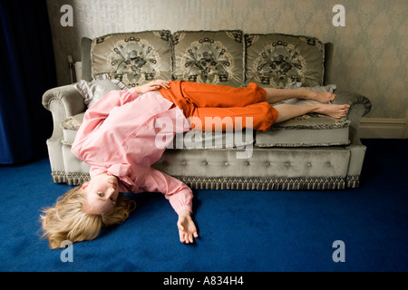 A blonde girl in casual dress lying inverted with her legs on a cozy sofa and head on the floor with blue carpet. Stock Photo