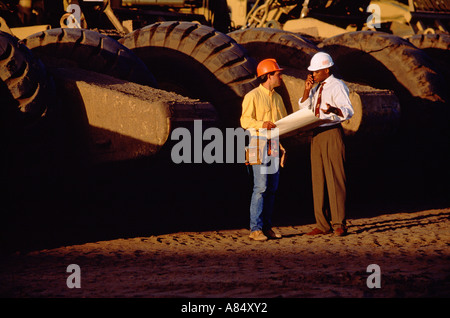 Two men standing outdoors by huge earth moving machine at road construction site. Stock Photo