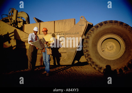 Two men standing outdoors by huge earth moving machine at road construction site. Stock Photo