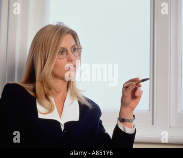 Young blonde executive woman sitting indoors in office, looking aside and pointing with pen. Stock Photo
