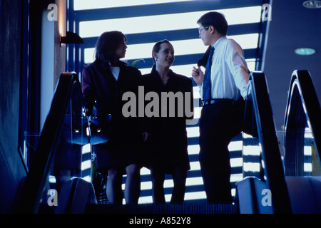 Three office workers in building standing at top of escalators. Stock Photo