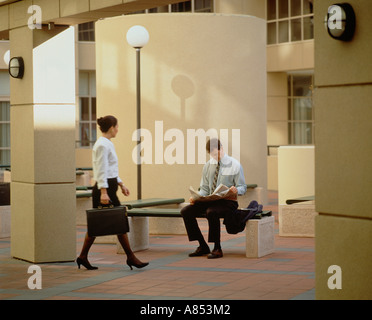 Young woman glancing at man reading newspaper as she walks past him in city mall. Stock Photo
